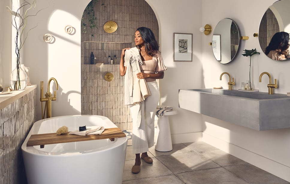 Organic luxe bathroom, brushed gold faucets, woman holding towel by tub.